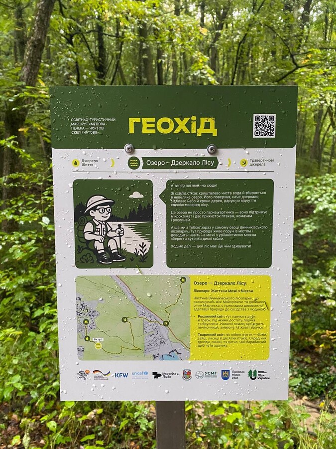 На фото - інформаційний стенд у лісі з написом &laquo;Геохід&raquo;. Табличка розповідає про природну локацію &laquo;Озеро &mdash; Дзеркало лісу&raquo; у межах Винниківського лісопарку. На ній подано карту маршруту, короткий опис місцевості, QR-код і логотипи партнерів проєкту.