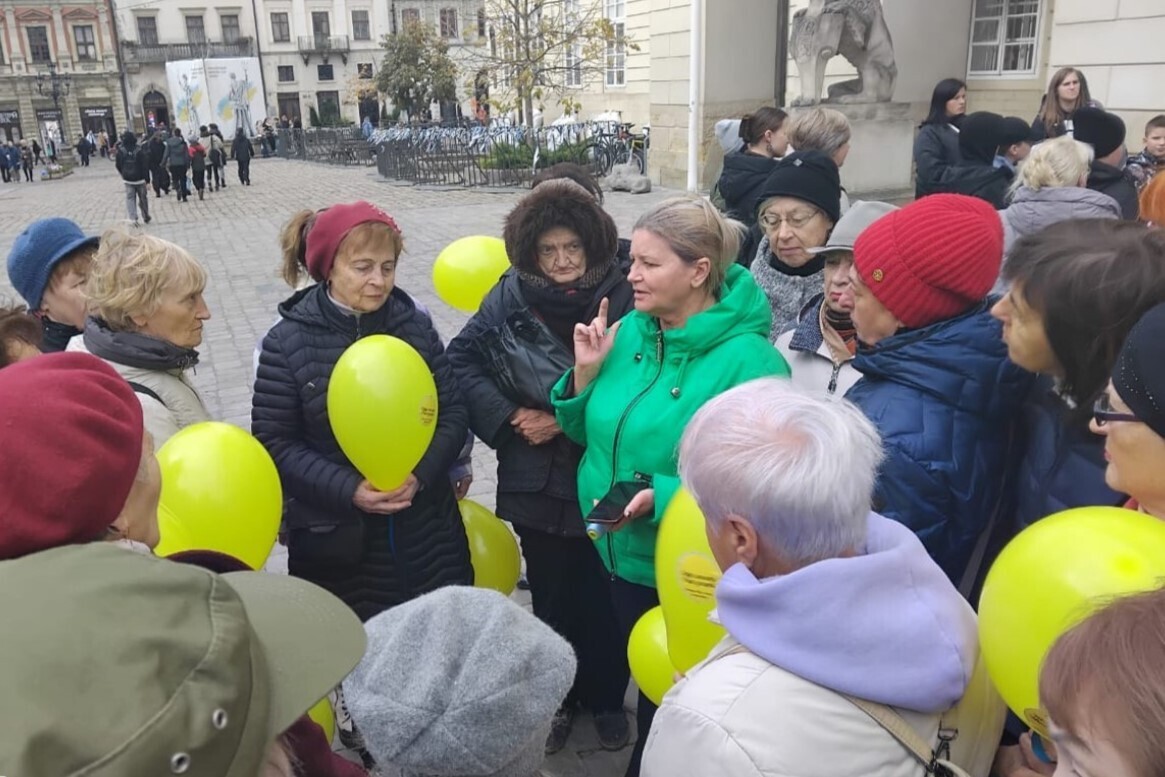 Люди літнього віку стоять на площі Ринок і слухають Людмилу Гришко, у руках тримають кульки жовтого кольору