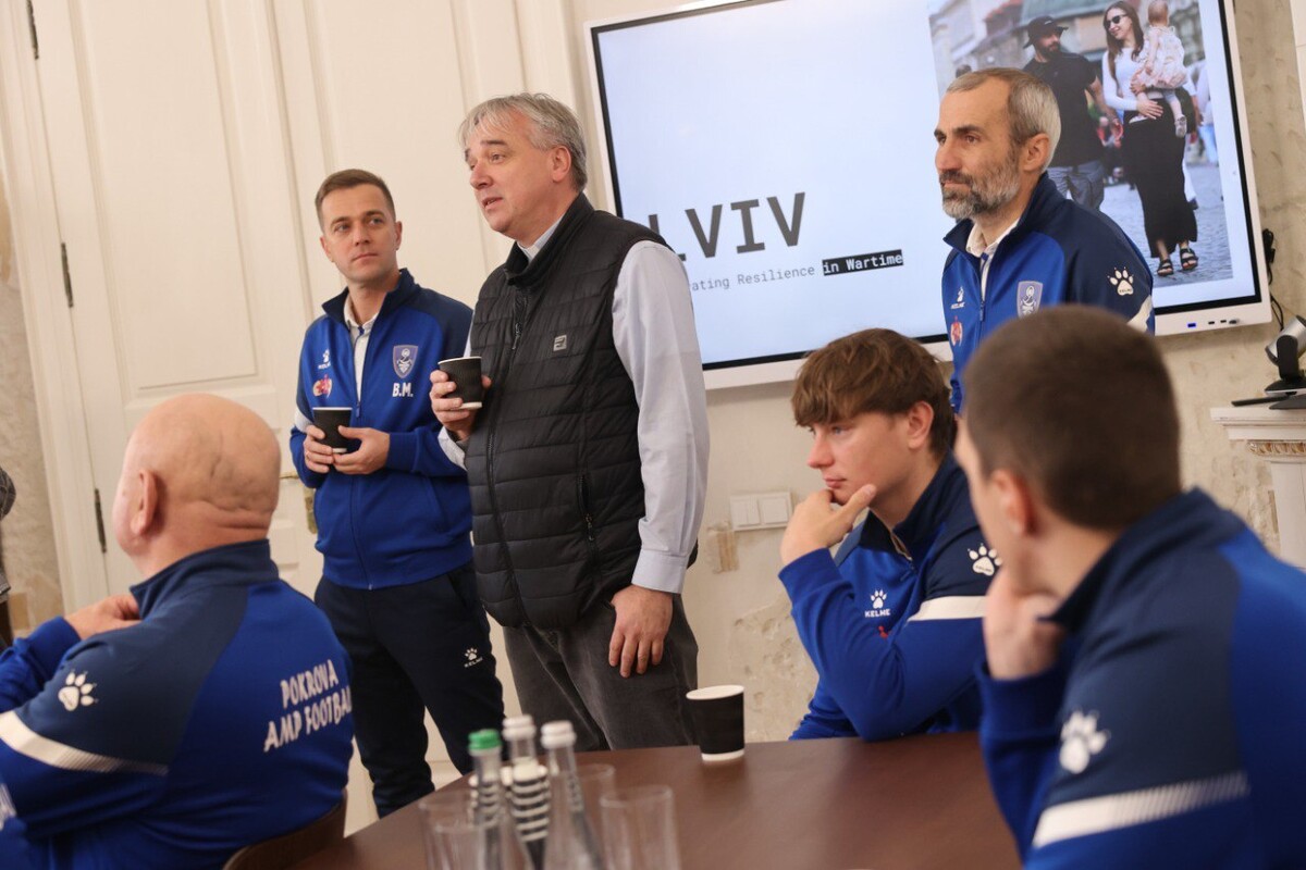На фото спортсмени та отець Михайло сидять і стоять у світлій кімнаті навколо столу. На екрані позаду показано слово &ldquo;LVIV&rdquo;.