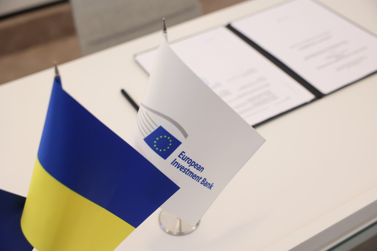 Маленькі прапорці України та Європейського інвестиційного банку стоять поруч на білому столі. На другому плані документи та ручка.