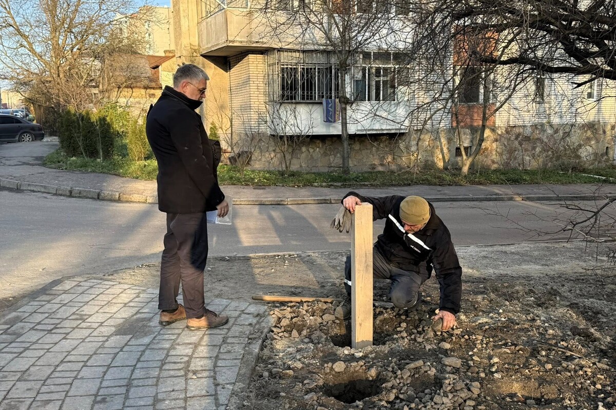 Чоловіки встановлюють дерев'яний стовпець в рамках робіт з ознакування теренкурів в парку Горіховий Гай. Один спостерігає за процесом, а інший присів біля викопаної ями й встановлює дерев'яний стовп. Навколо видно землю, каміння та інструмент - лопату.