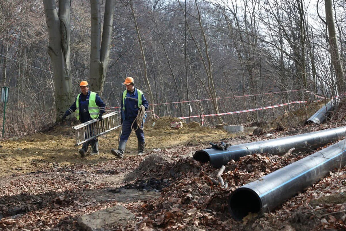 Два працівники Львівводоканалу, одягнені у спецогдяг &ndash; один несе драбину, інший мотузку, прямують вперед. Попереду чоловіків лежать поліетиленові труби, а позаду росте багато дерев