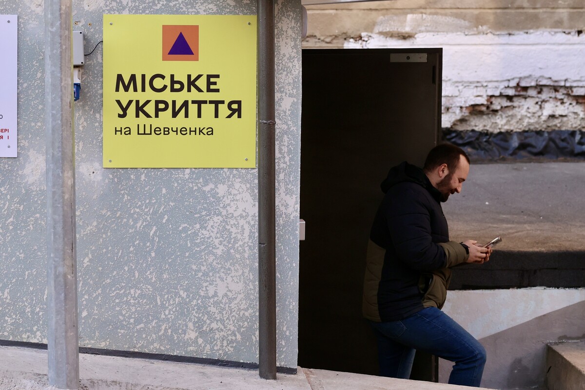 Чоловік виходить з укриття, дивлячись у свій смартфон, поруч на стіні жовта табличка з написом &laquo;МІСЬКЕ УКРИТТЯ на Шевченка&raquo;.