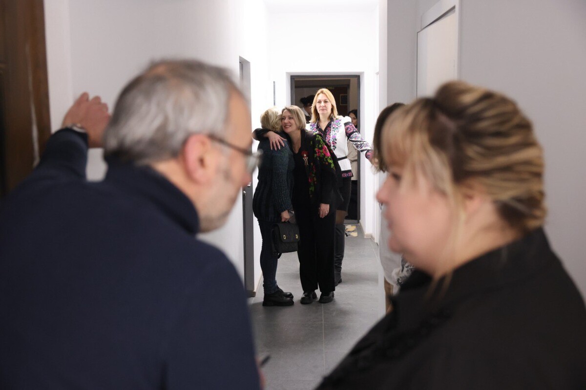 На передньому плані двоє людей спілкуються в коридорі простору. Позаду - троє жінок: одна обіймає іншу, третя спостерігає поруч.
