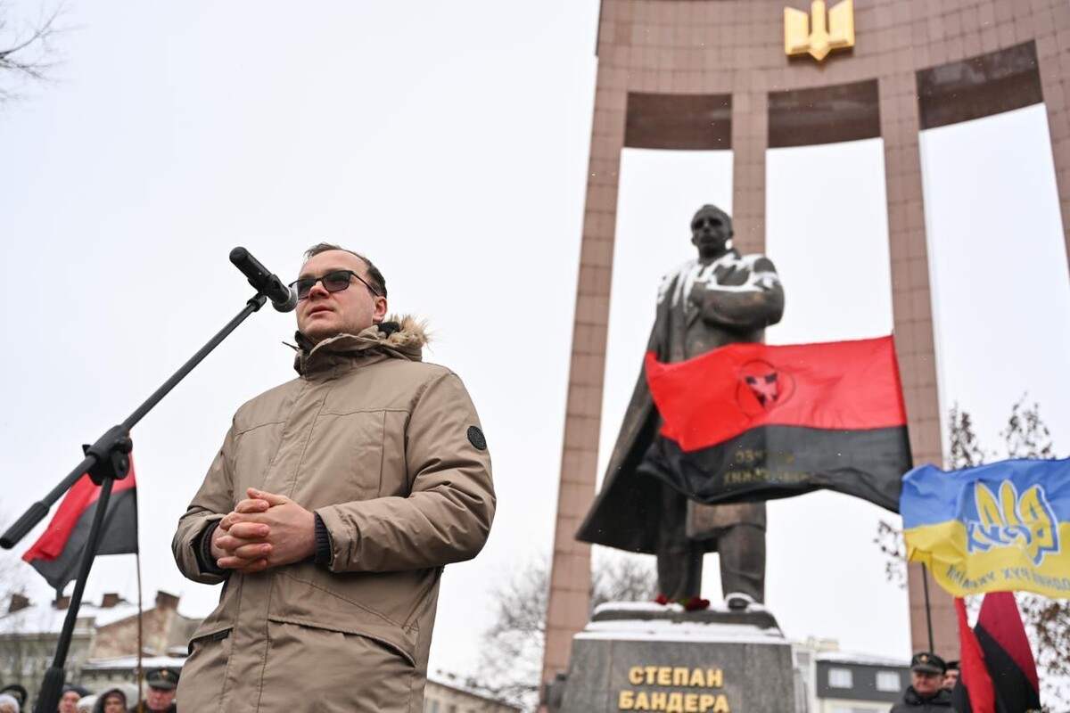 Перший заступник міського голови Андрій Москаленко говорить у мікрофон. Позаду видно червоно-чорний та синьо-жовтий прапори і пам&rsquo;ятник Степану Бандері.