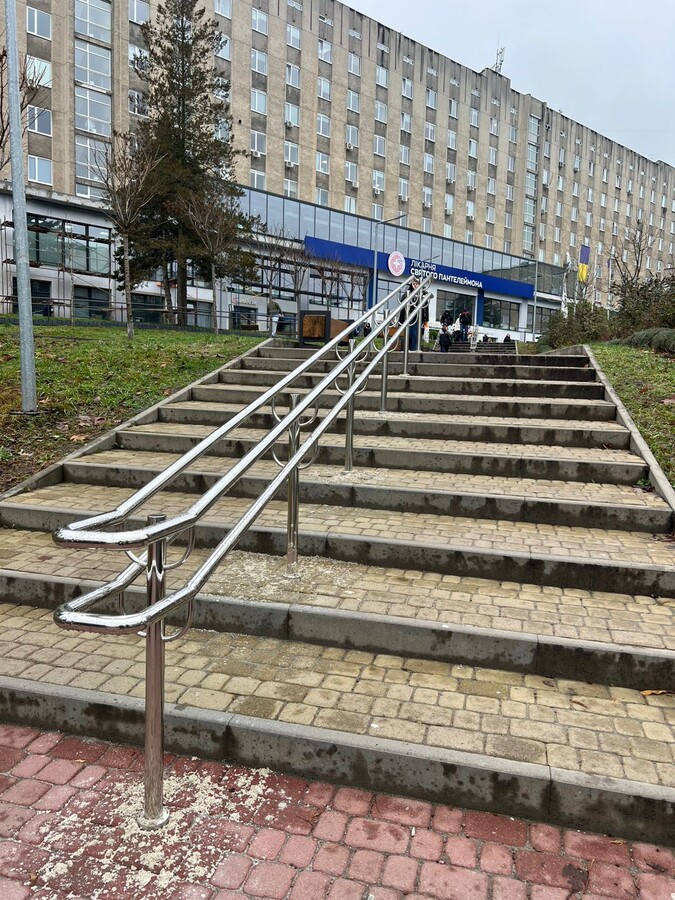 Довгі срібні поручні, біля яких з обох боків сходи вимощені бруківкою. На фоні велика світла будівля - лікарня Св. Пантелеймона