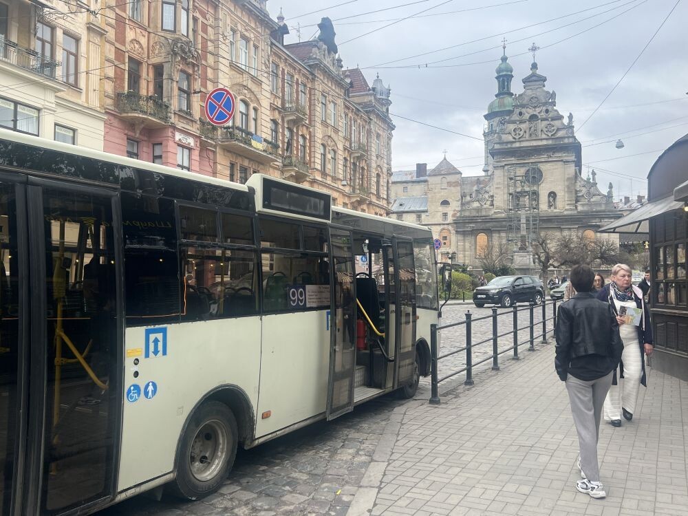 Автобус білого кольору з написом № 99 стоїть на зупинці із відчиненими дверима. Поруч проходять люди