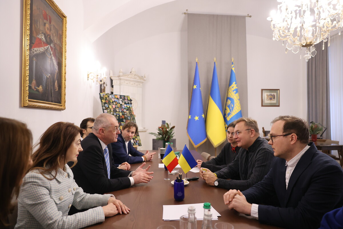 Представники двох сторін сидять за столом. Позаду видно три прапори: України, Європейського Союзу та синьо-жовтий прапор із гербом Львова. На столі також два прапори: України та Румунії.