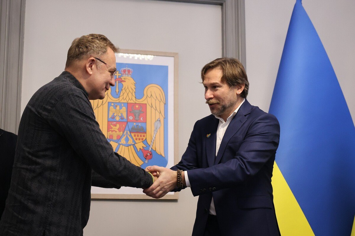 Почесний консул Ростислав Вовк тисне руку меру Львова Андрію Садовому. Позаду видно синьо-жовтий прапор України та державний герб Румунії.