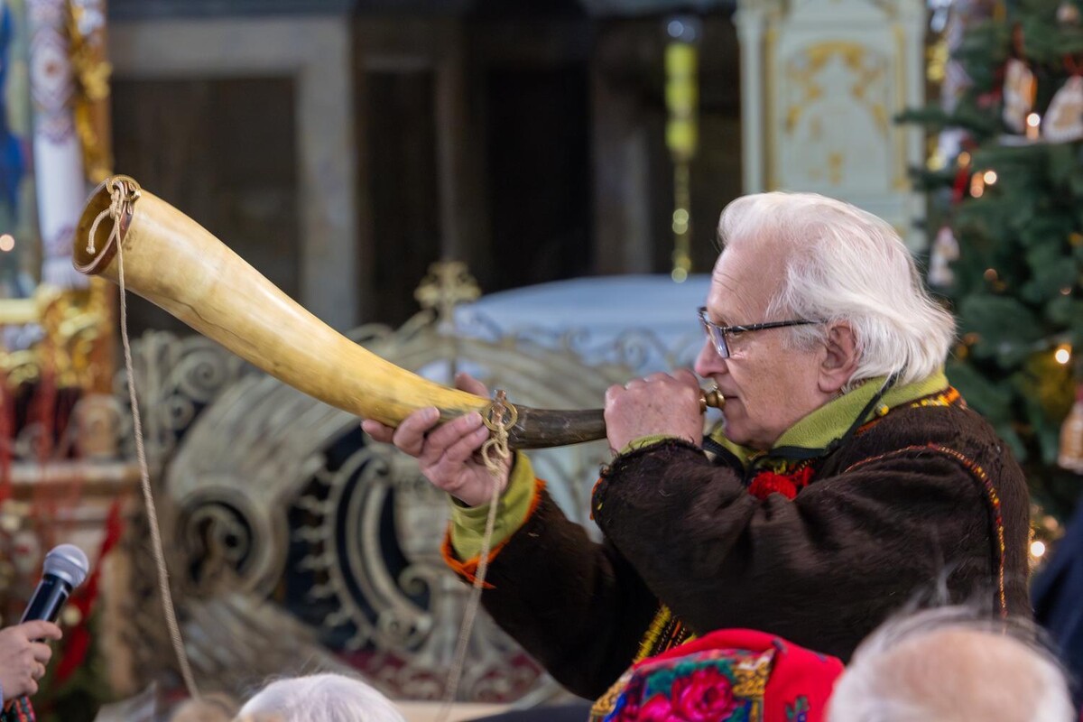 Чоловік одягнений у традиційний народний стрій темних і зелених відтінків стоїть у храмі та сурмить у ріжок, духовий народний інструмент, тримаючи його обома руками.