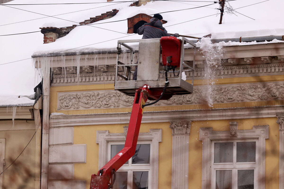 Фасад будівлі жовтого кольору з білими декоративними елементами навколо вікон і під дахом. Будинок має архітектурні прикраси &mdash; ліпнину з рослинними візерунками. На даху лежить товстий шар снігу, а з краю даху звисають довгі бурульки. Біля даху працює чоловік у темному зимовому одязі та шапці. Він стоїть у металевій підйомній люльці, закріпленій на червоному автопідіймачі. Люлька піднята на рівень даху. Чоловік лопатою скидає сніг із краю покрівлі.