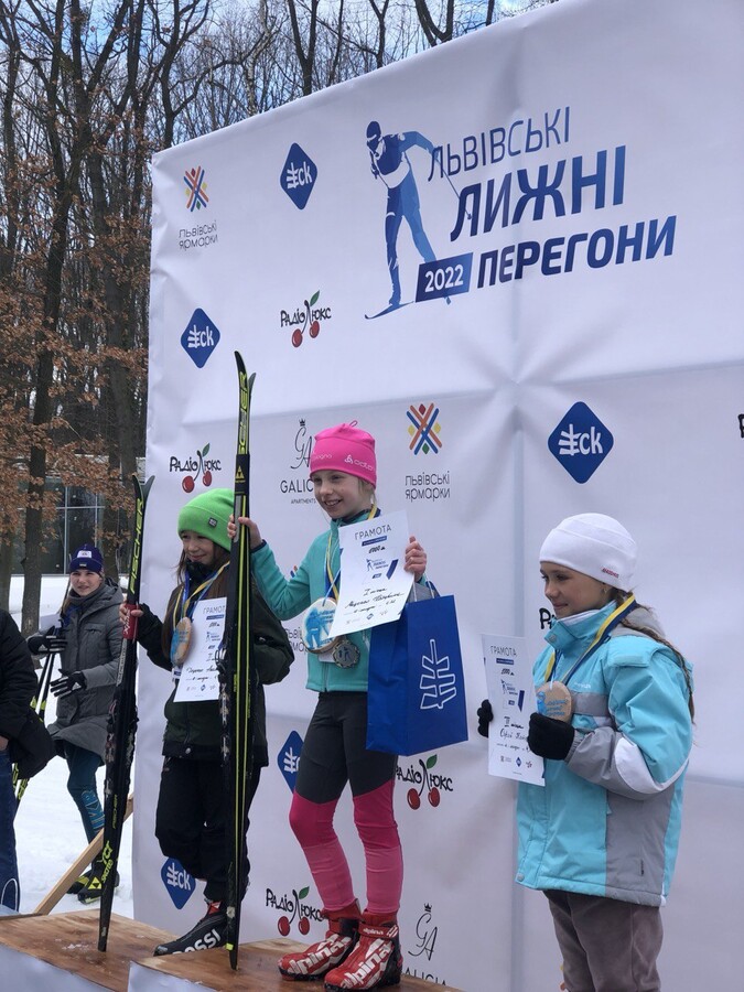 Нагородження юних учасниць лижних перегонів. Троє дівчат стоять на п&rsquo;єдесталі пошани з лижами в руках. На їхніх шиях &mdash; медалі, а в руках &mdash; грамоти та подарункові пакети. Усі вони одягнені в теплий спортивний одяг і зимові шапки. Позаду &mdash; брендована фотозона з написом &laquo;Львівські лижні перегони 2022&raquo; та логотипами партнерів заходу.