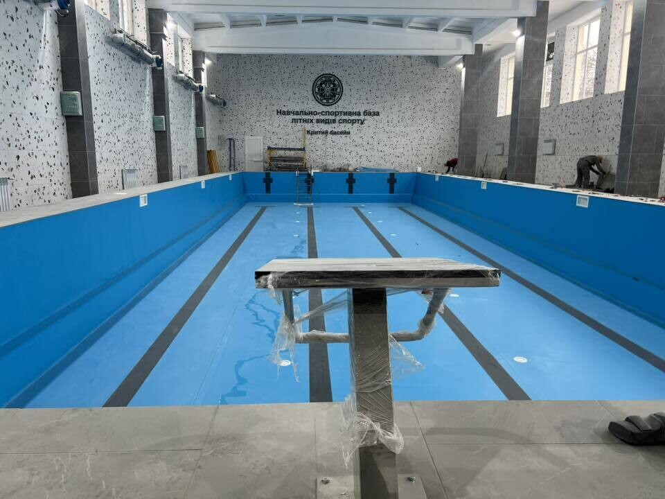 Зблизька зображено стартову тумбу, обгорнуту захисною плівкою. Позаду яскраво-блакитна чаша з чорними смугами розмітки, що веде до протилежної стіни з написом &laquo;Навчально-спортивна база літніх видів спорту&raquo;.