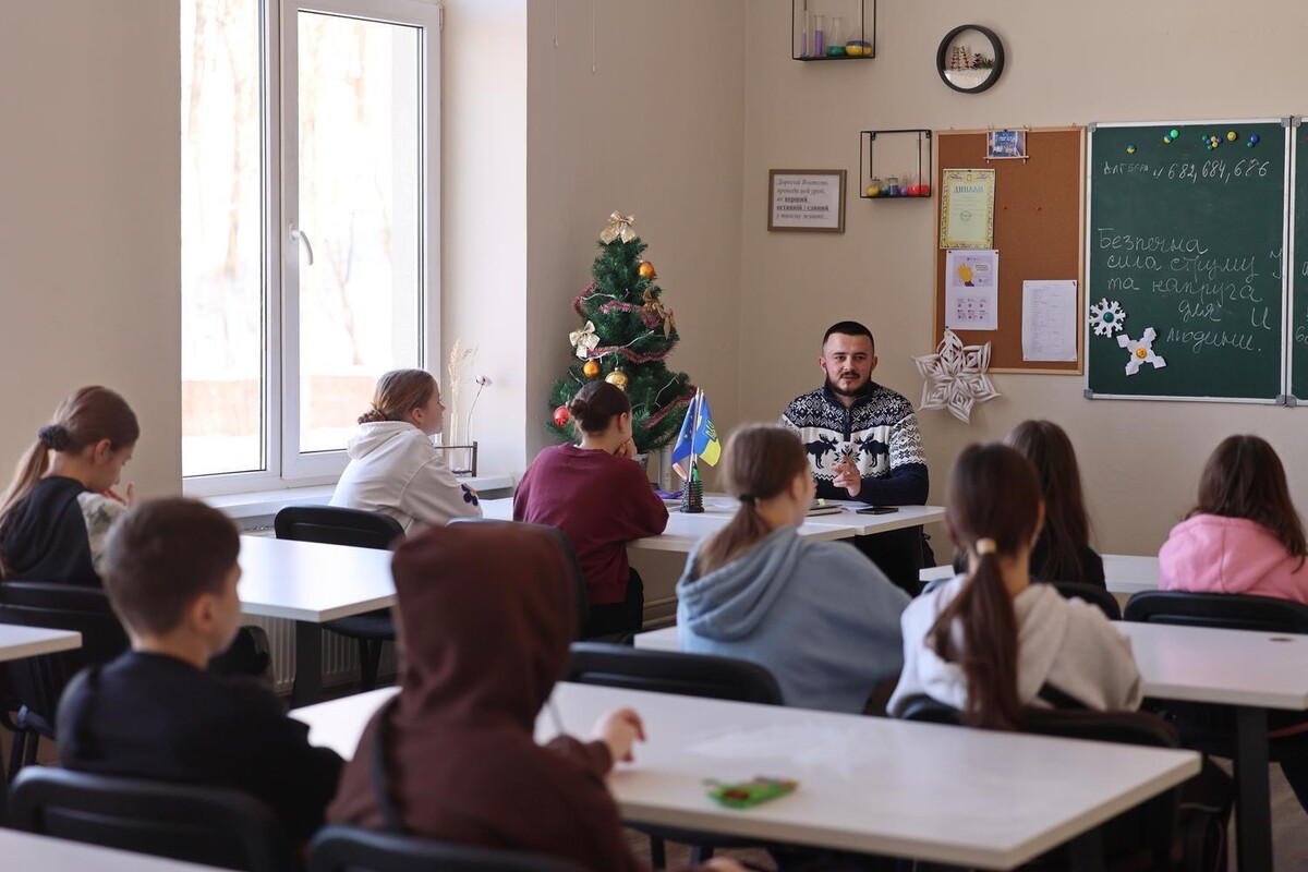 Ігор Дусан сидить за вчительським столом у класі, де за партами сидять учні.