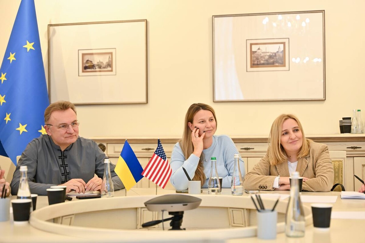 Зліва мер Львова Андрій Садовий, по центру депутатка Львівської міськради Вікторія Христенко, а справа депутатка Львівської міськради та очільниця Інституту стратегії культури Юлія Хомчин за столом у Білій залі Львівської міської ради під час онлайн-зустрічі з представниками міста Тампа у США. Позаду прапори Львова, України та ЄС. На стінах картини міста. На столі два прапорці України та США.