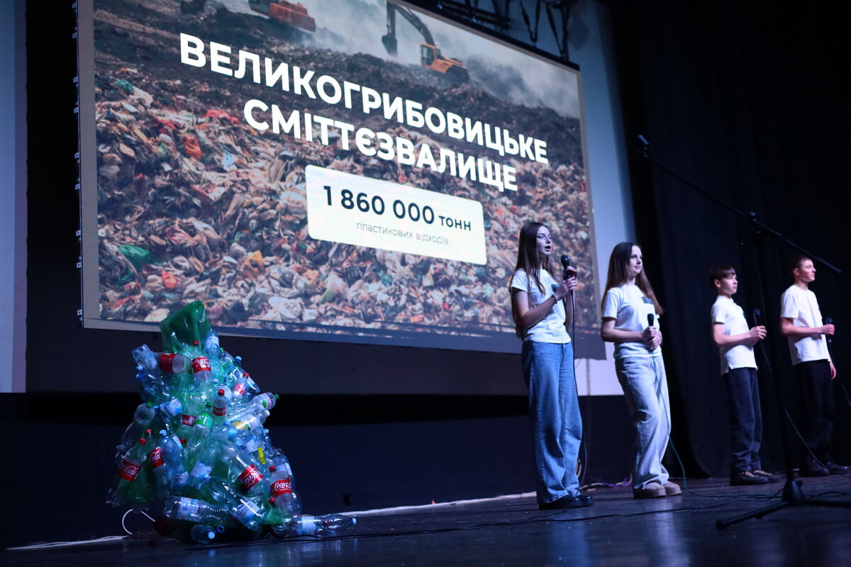 Учні стоять на сцені та говорять у мікрофон. Позаду на екрані напис “Великогрибовицьке сміттєзвалище 1 860 000 тонн пластикових відходів.
