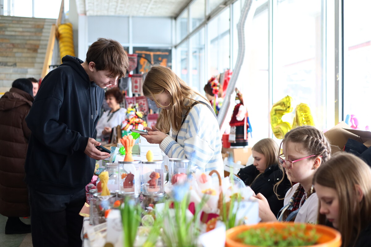 Школярі продають власноруч зроблені вироби за столом. Поруч стоять відвідувачі, які розглядають товари та спілкуються з продавцями.