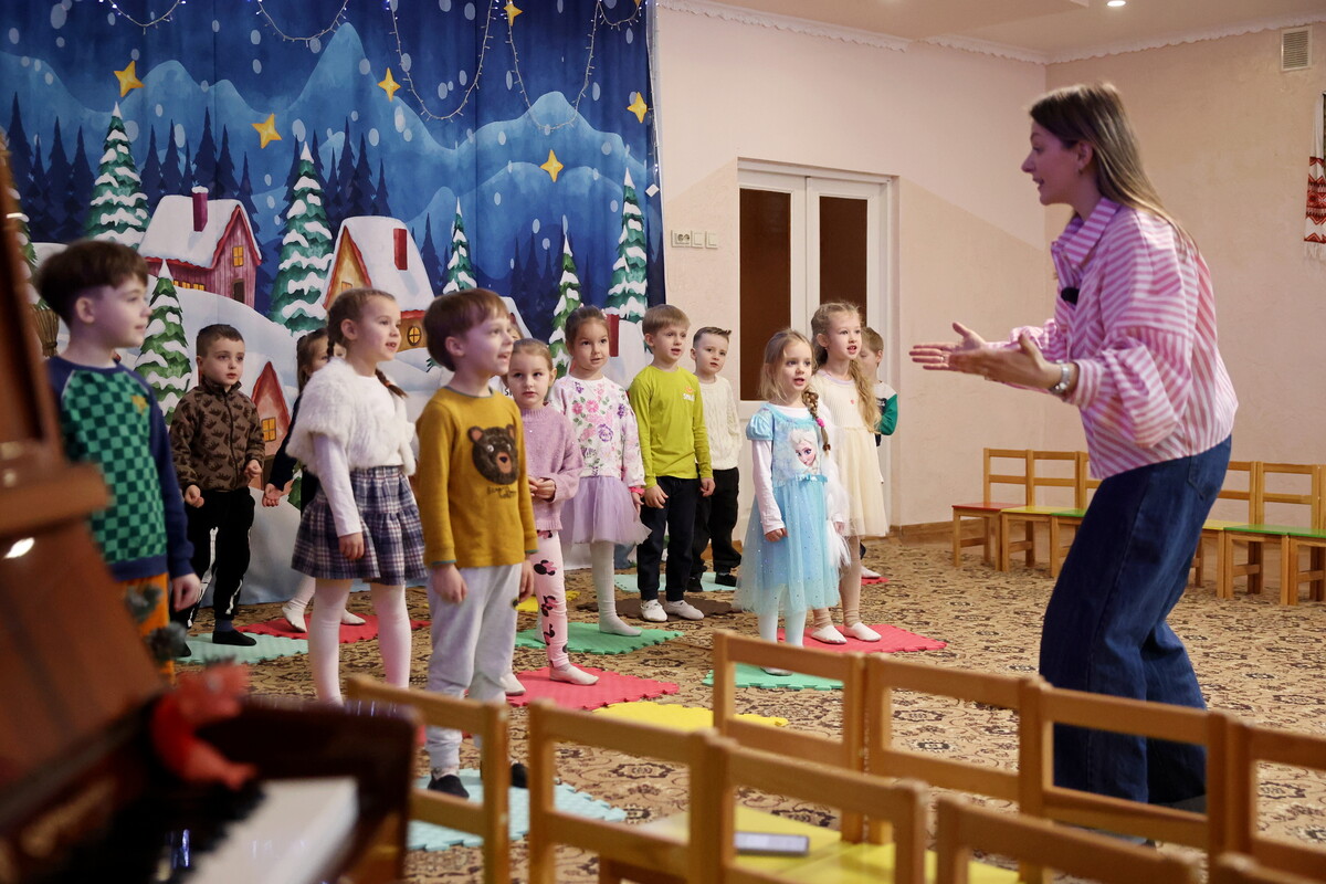 Устина показує рухи дітям у концертному залі садка на тлі декорацій із засніженими будиночками.