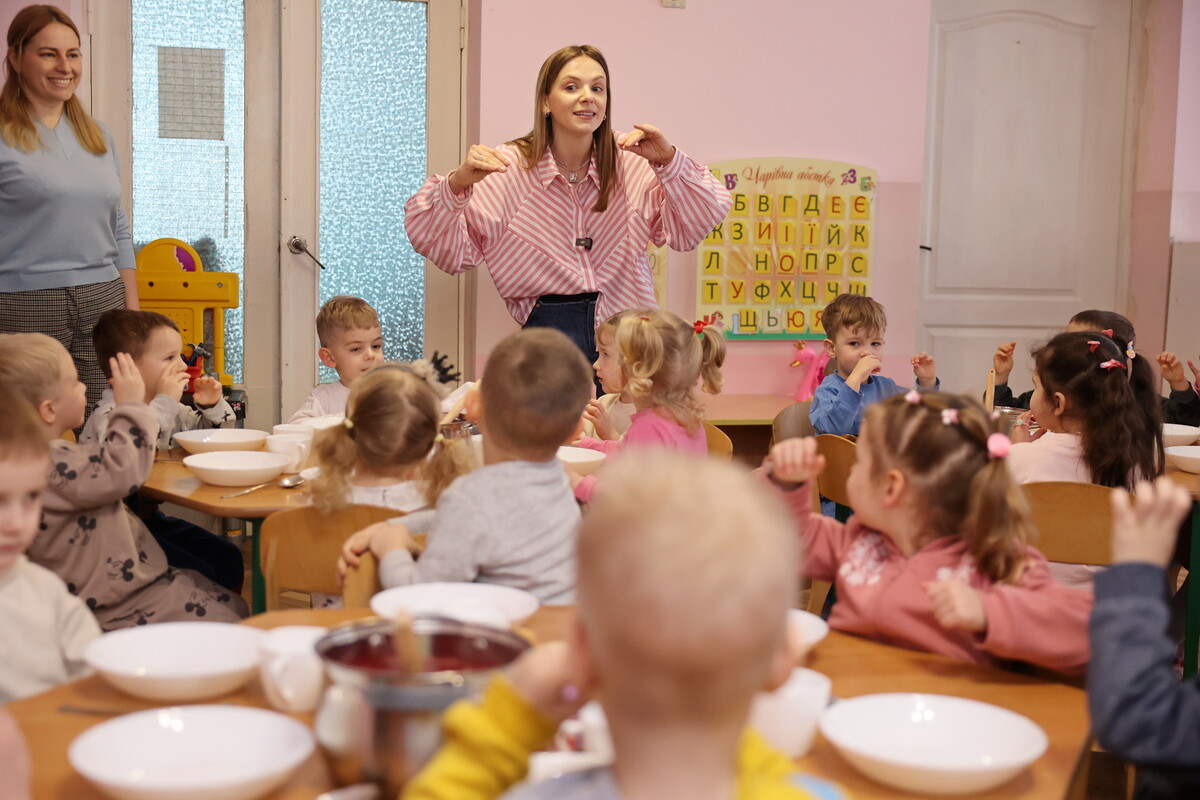 Діти, які готуються до обіду, сидять за столами з тарілками, спостерігаючи за Устиною та повторюючи за нею рухи.