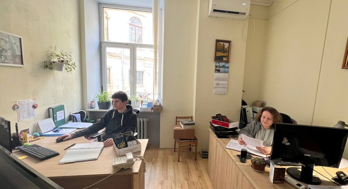 Двоє працівників Бюро знахідок, чоловік та жінка, працюють за робочими столами. На стінах картини, висить календар та кондиціонер. Позаду них вікно. На столах документи, журнали та компʼютери
