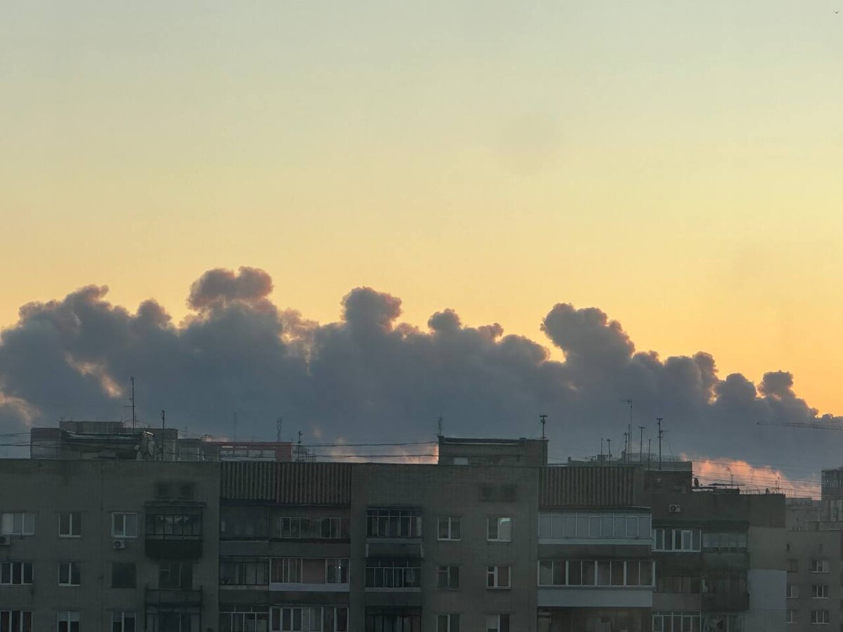 На передньому плані &mdash; багатоповерхові житлові будинки, позаду них в небо підіймаються густі темно-сірі хмари диму. Це наслідок ворожого влучання десь неподалік