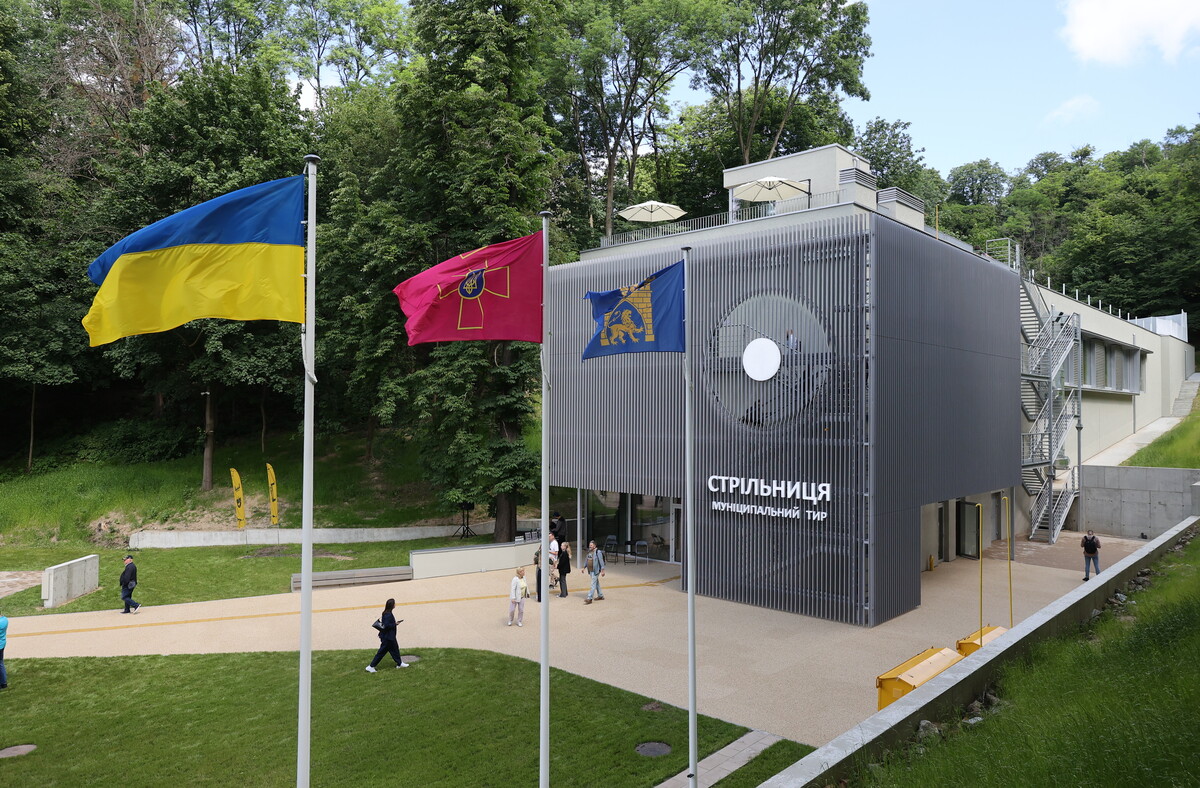 Муніципальний тир "Стрільниця": велика модерна будівля сірого кольору. Поруч на флагштоках синьо-жовтий прапор, прапор Збройних сил України та прапор Львова