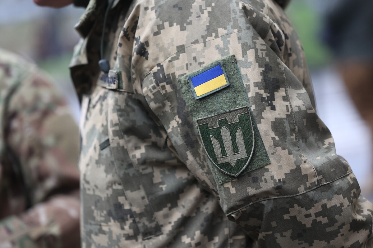 Ілюстративне фото - плече чоловіка у військовій формі із шевроном та маленьким прапором України на ньому