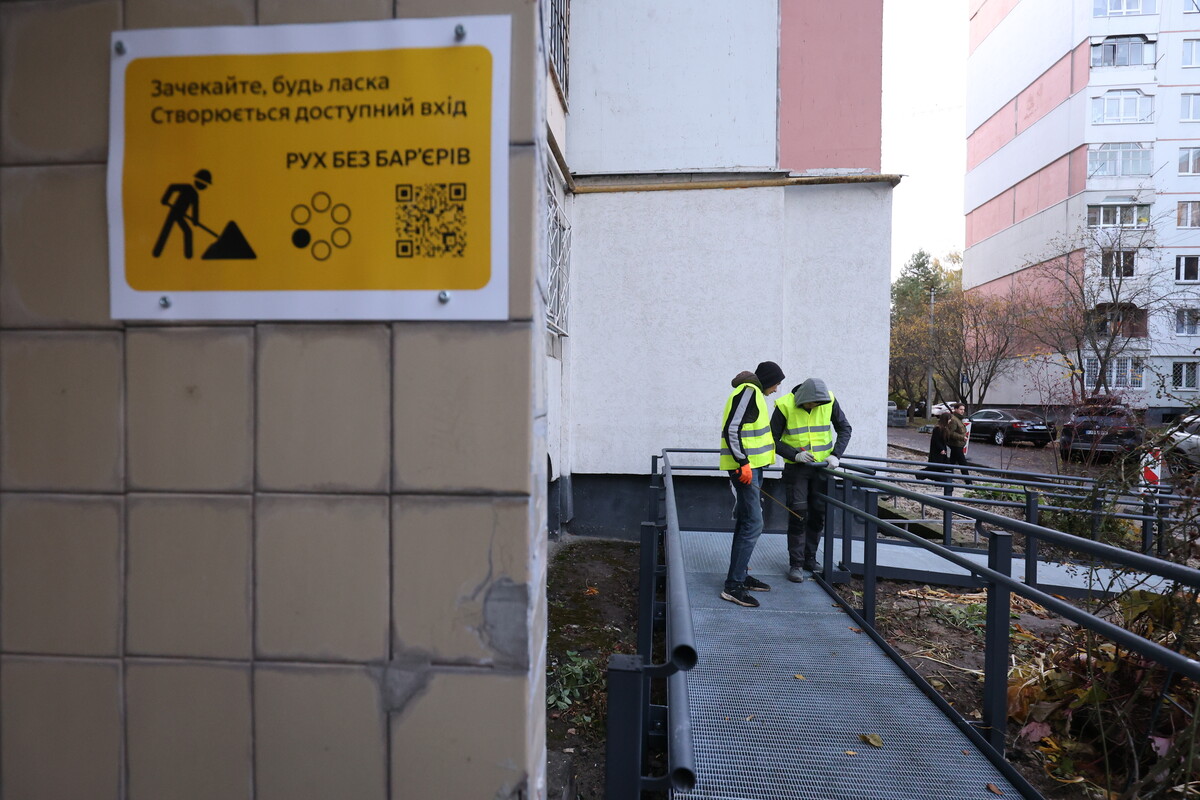 На фото зображено вхід до житлового будинку, біля якого облаштовують пандус для людей з інвалідністю. Ліворуч на стіні будівлі висить жовта інформаційна табличка. На ній українською написано: &laquo;Зачекайте, будь ласка. Створюється доступний вхід. Рух без бар&rsquo;єрів&raquo;.