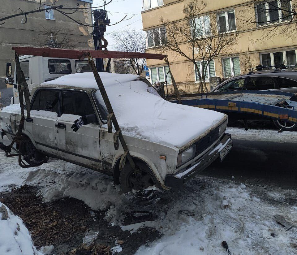 На засніженій вулиці на тлі житлових багатоповерхівок евакуатор за допомогою спеціальних кріплень підіймає вкритий шаром снігу легковий автомобіль білого кольору