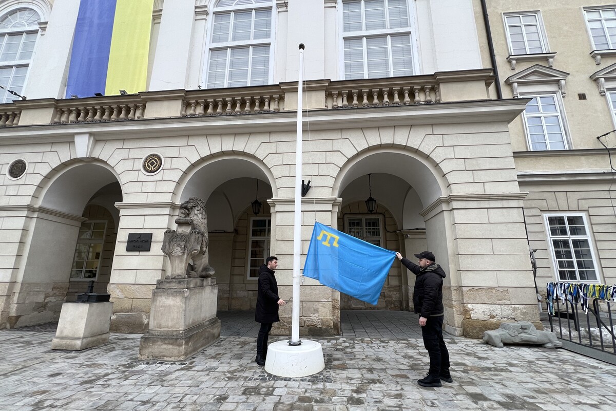 Момент підняття прапора: біля флагштоку перед будівлею міської Ратуші двоє чоловіків у темному одязі піднімають кримськотатарський прапор: один із них тримає прапор, а інший допомагає його підняти. Ліворуч біля входу є кам&rsquo;яна скульптура лева з гербом, праворуч &ndash; огорожа з прив&rsquo;язаними синьо-жовтими стрічками