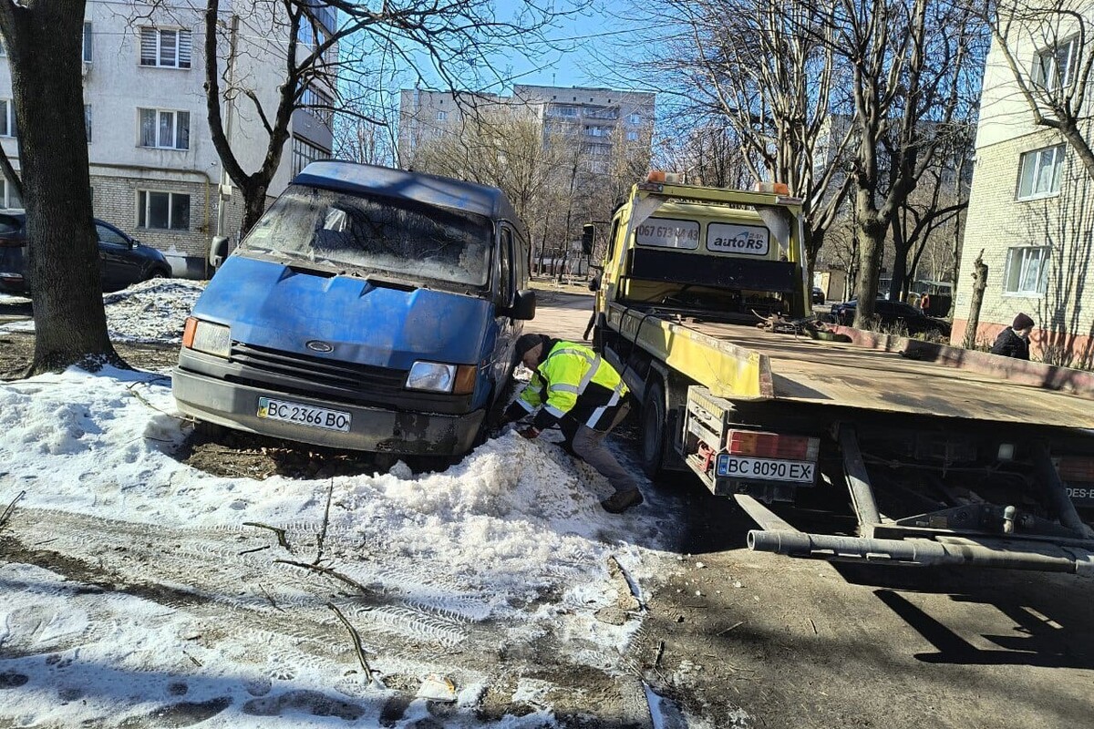 Покинутий синій мікроавтобус. Поруч з ним стоїть евакуатор. Працівник у спецодязі схилився до колеса покинутого автомобіля. Довкола машини - сніг. Поруч багатоповерхові житлові будинки та дерева між ними.