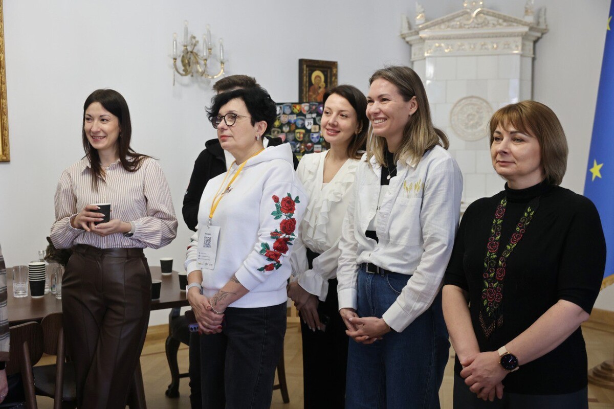 Учасники резиденції, у кабінеті мера Львова слухають презентацію та усміхаються. Позаду них грубка вкрита білим кахелем. Зліва частково видно прапор ЄС