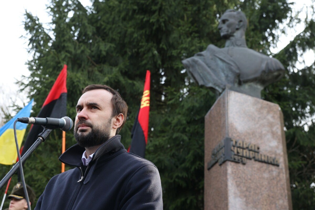Керуючий справами виконкому Львівської міськради Євген Бойко під час виступу на вшануванні річниці загибелі Романа Шухевича перед пам&rsquo;ятником генерал-хорунжого УПА на Білогорщі. Позаду посадовця височіє металеве погруддя Шухевича, а на гранітній плиті напис його псевдо &laquo;Тарас Чупринка&raquo;. За пам&rsquo;ятником росте велика зелена ялина