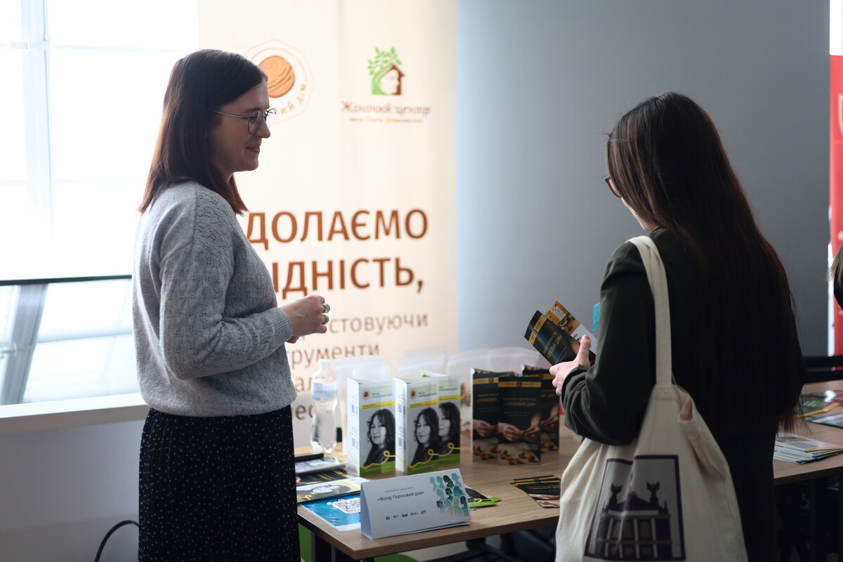 На ярмарку жіночих правозахисних організацій біля інформаційного стенда стоять дві жінки. Зліва жінка в окулярах та світлому светрі дивиться на співрозмовницю, яка тримає в руках буклет. На столі перед ними розкладені візитівки та брошури, а на другому плані видно великий банер із написом &ldquo;Долаємо бідність&rdquo;