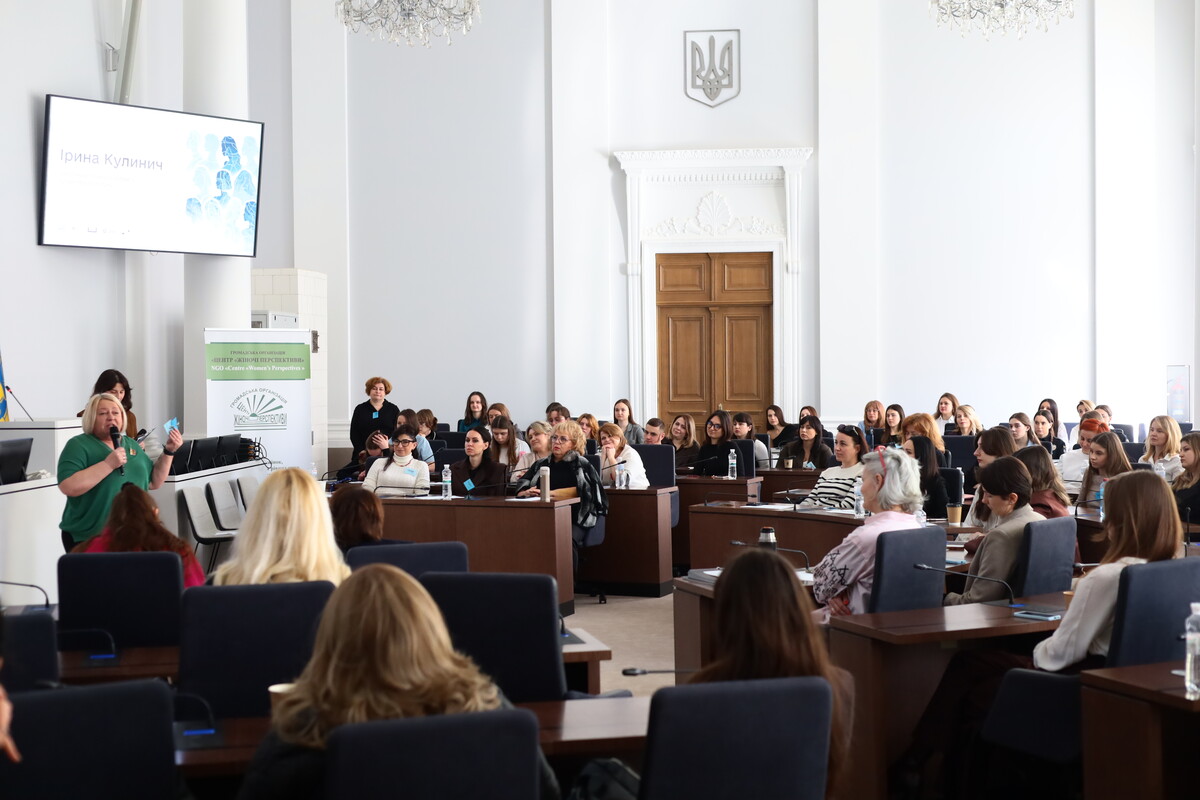 У сесійній залі за столами сидять учасниці заходу, перед ними виступає заступниця міського голови Львова Ірина Кулинич. На великому екрані позаду неї відображено її ім&rsquo;я та прізвище на світлому тлі