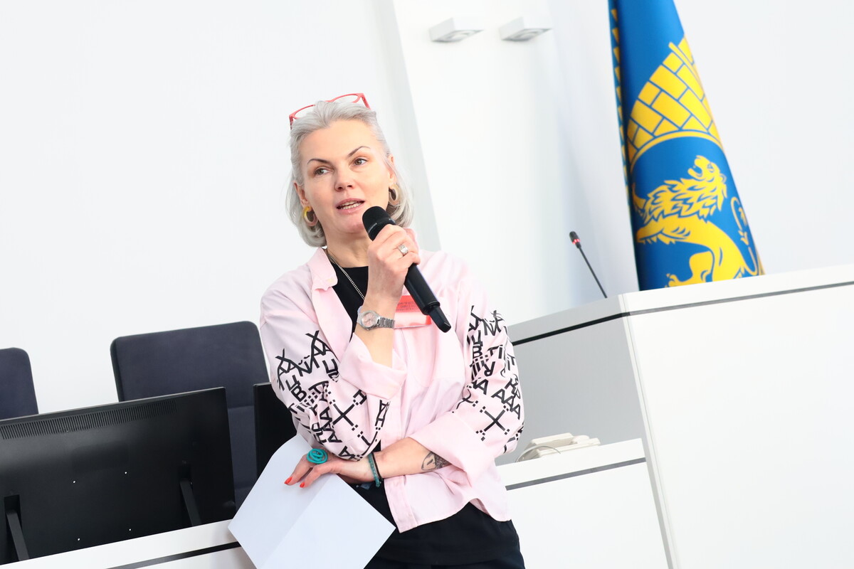 Мистецтвознавиця та соціальна працівниця центру НЕЗЛАМНІ Катерина Кіт-Садова виступає з мікрофоном у руках, а в іншій руці тримає аркуш паперу. На другому плані праворуч, біля білої стіни, видно прапор міста Львова