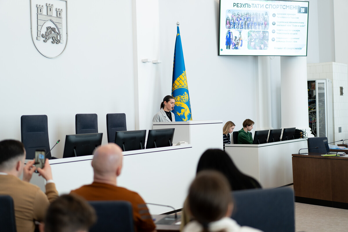 Презентація тренерських досягнень у великій залі засідань Львівської міської ради. На білій стіні висить герб і прапор Львові. Тренерка стоїть за трибуною та доповідає про результати спортсменів, презентація яких транслюється на великому екрані праворуч. У залі також присутні представники комісії та інші тренери.