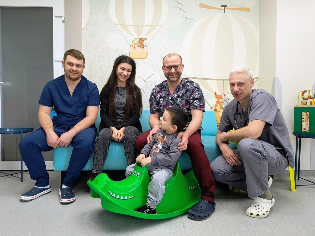 Хлопчик усміхається сидить верхи на зеленій іграшці-гойдалці у формі крокодила. Позаду нього на дивані сидить мати та медики лікарні святого Миколая Першого медоб’єднання Львова