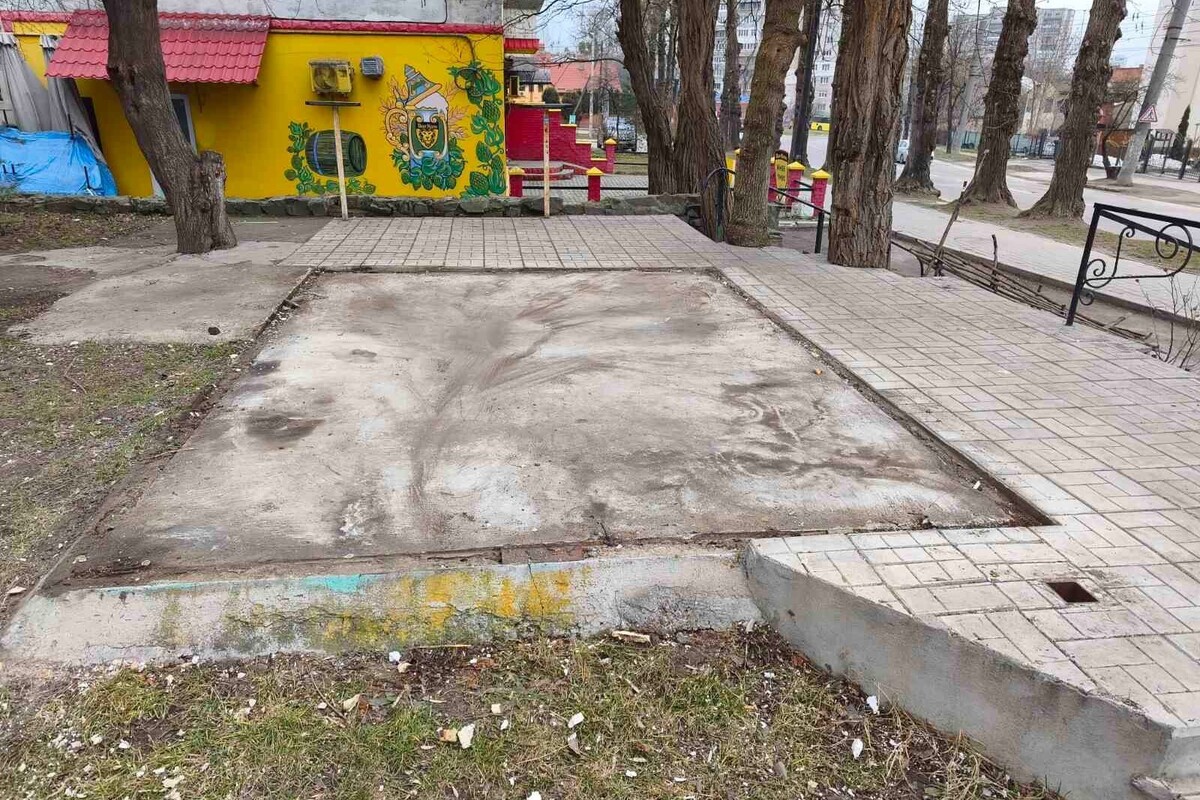 На тротуарі видно місце, де раніше стояв кіоск. Поруч частина будівлі з розмальованим фасадом жовтого кольору. Довкола ростуть дерева