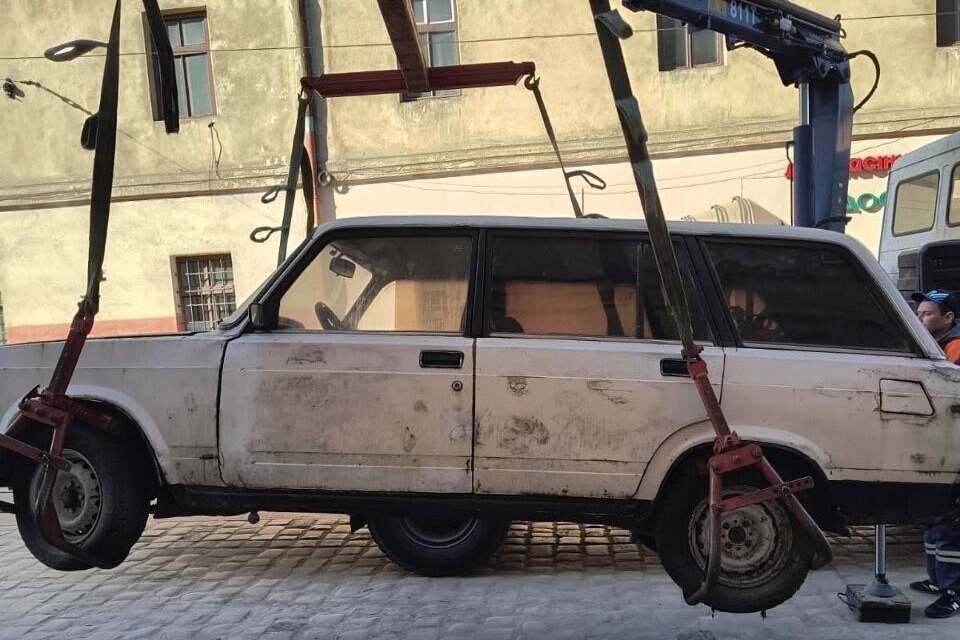 Евакуатор за допомогою крана-маніпулятора підійняв у повітря білий старий автомобіль, закріпивши його спеціальними захватами за колеса. Машина висить над брукованою дорогою на тлі будинку