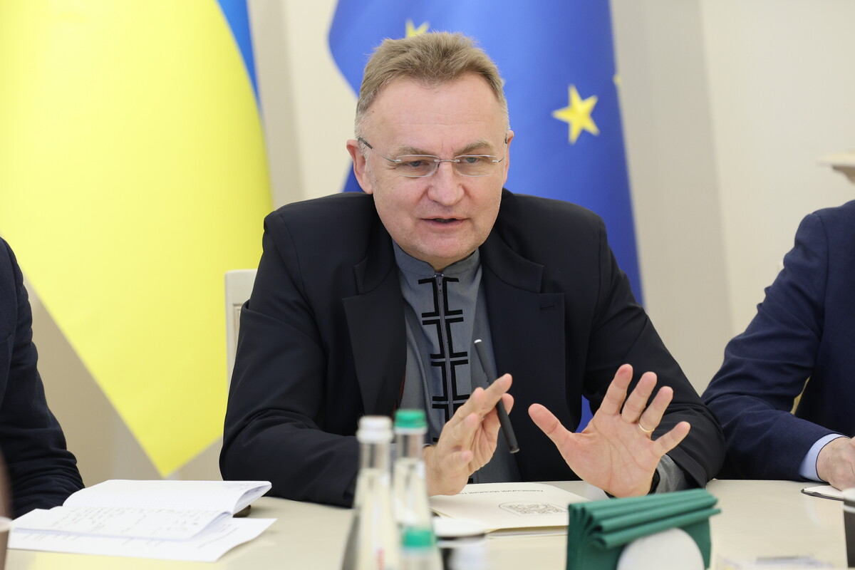 Андрій Садовий, який сидить за круглим столом у мерії на фоні прапорів України та ЄС. Мер Львова активно жестикулює руками, пояснюючи свою думку, а перед ним на столі лежать розгорнуті блокноти з нотатками. Він одягнений у темний піджак та сіру сорочку з вишитим чорним орнаментом.