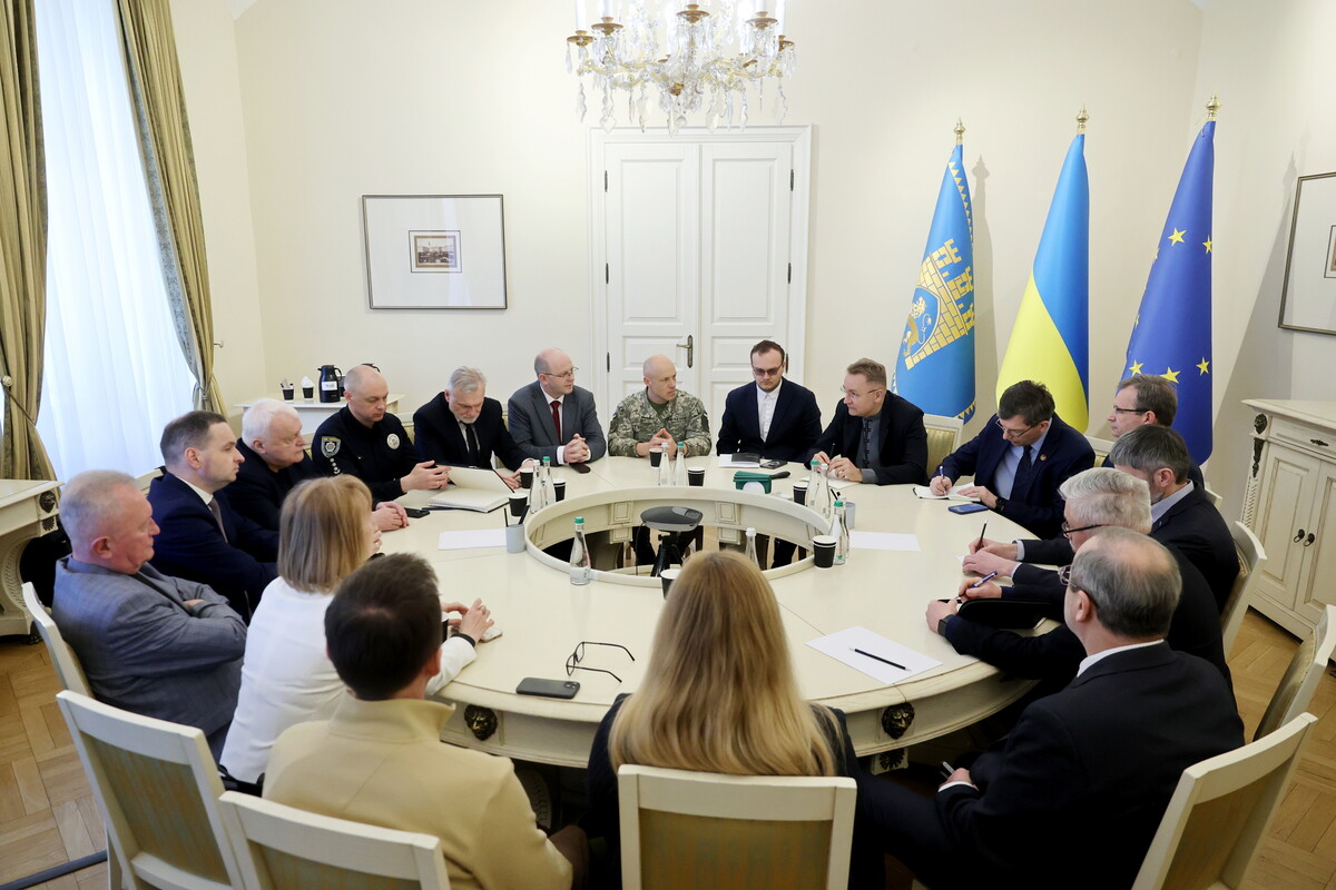 Загальний план великої наради у світлій залі мерії з високою кришталевою люстрою. Близько п&rsquo;ятнадцяти осіб &mdash; ректори львівських університетів, представники поліції та міської влади &mdash; сидять навколо великого білого круглого столу. На столі стоять паперові стаканчики з кавою, пляшки води, блокноти для записів та мобільні телефони. Учасники наради уважно слухають Андрія Садового, який сидить у правій частині кадру.