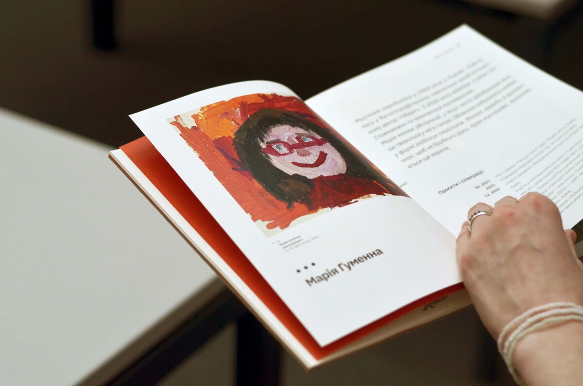 Розгорнута книга &ldquo;Art Brut&rdquo;, яку тримає рука людини. На лівій сторінці розміщено яскравий портрет дівчини в окулярах, виконаний у помаранчевих та червоних кольорах, а під ним написано ім'я &mdash; Марія Гуменна. На правій сторінці - текст українською мовою та перелік проєктів художниці.