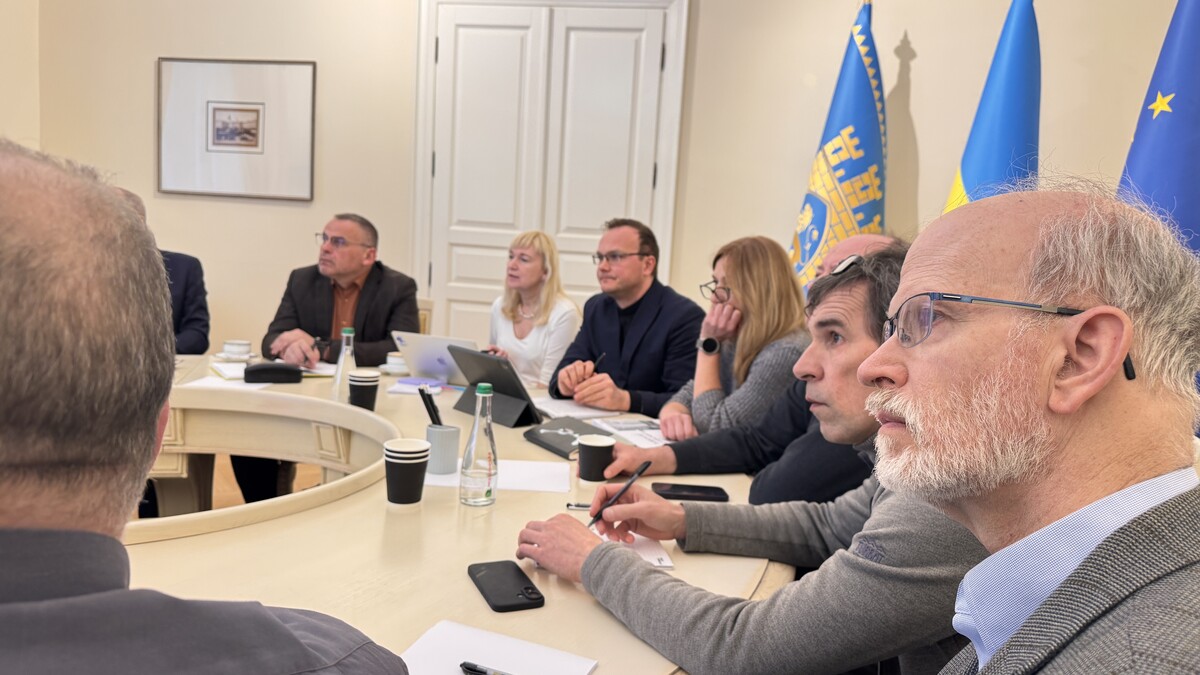 Члени робочої групи щодо розвитку території Львівського автобусного заводу, посадовці міськради, представники виробництв та університетів міста за круглим столом у Білій залі Львівської міської ради під час першого засідання робочої групи. Усі дивляться на екран. Позаду прапори ЄС, України та міста