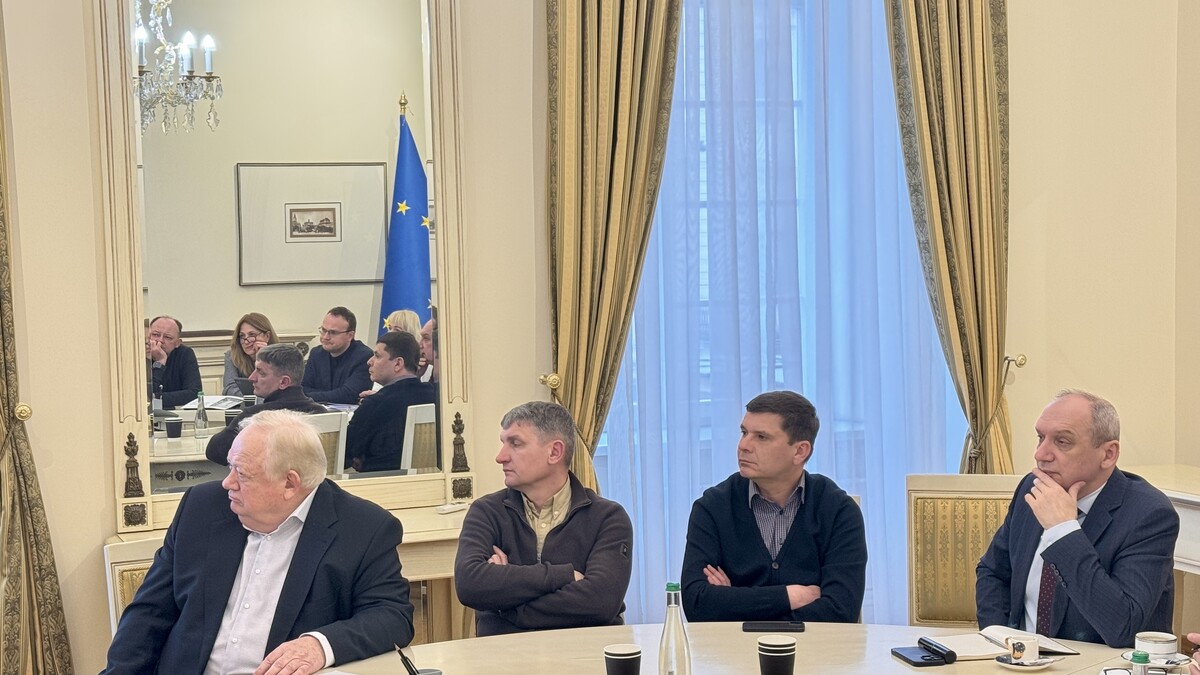 Четверо чоловіків, учасників робочої групи щодо розвитку території Львівського автобусного заводу, сидять за столом та дивляться на презентацію. У відображенні дзеркала за ними видно інших учасників робочої групи та частину прапора ЄС