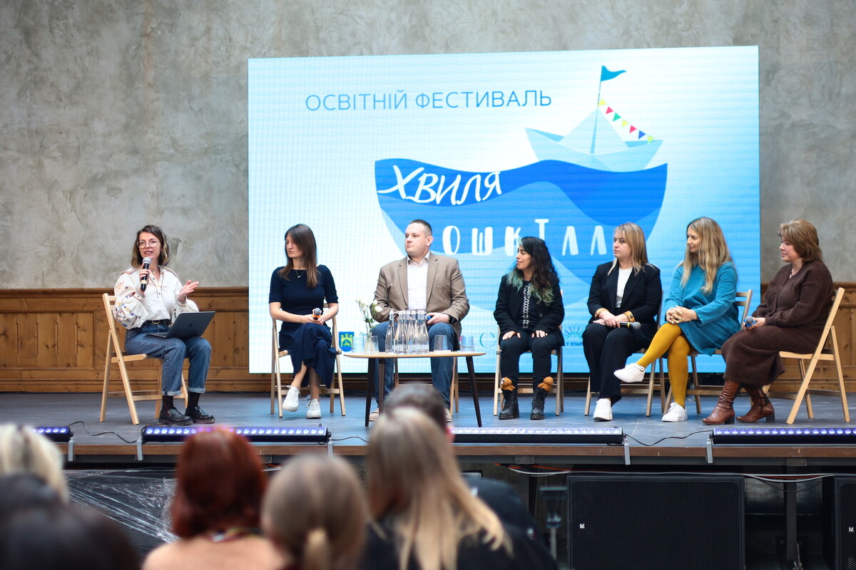 Семеро спікерів сидять на сцені під час дискусії. Одна з доповідачок розповідає, решта - слухають. Позаду спікерів на сцені великий екран зі зображенням паперового кораблика та хвиль. Напис: "Освітній фестиваль. Хвиля дошкілля"