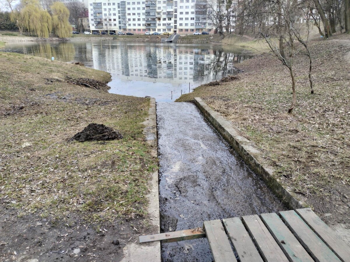 Невелике озеро зі спокійною водою, на протилежному березі багатоповерхівка. До води веде вузький бетонний спуск або канал, наповнений темною водою та мулом &mdash; ймовірно, це місце для стоку або очищення.