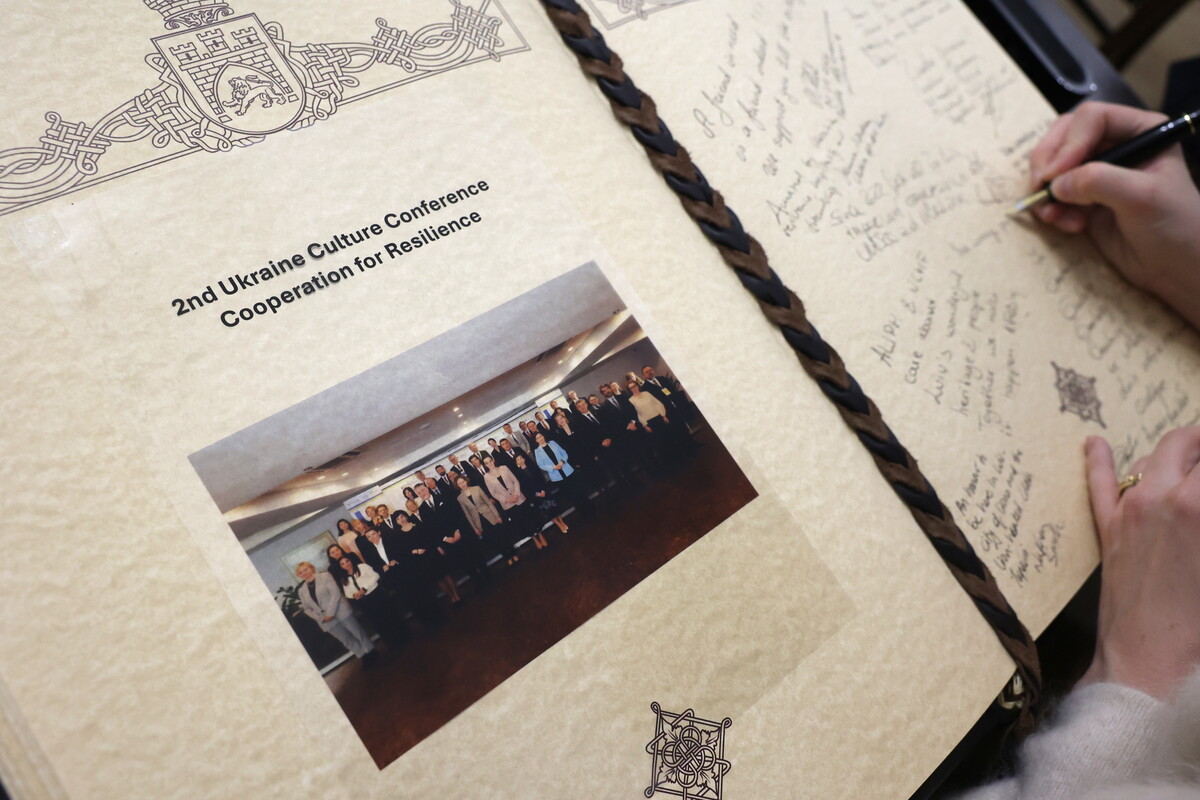 Відкрита сторінка Книги почесних гостей міста Львова з декоративним візерунком у кутах та зображенням герба. На лівій сторінці вклеєно групову кольорову фотографію учасників делегацій. На правій сторінці видно записи та підписи різними мовами, а хтось продовжує писати ручкою у Книзі.