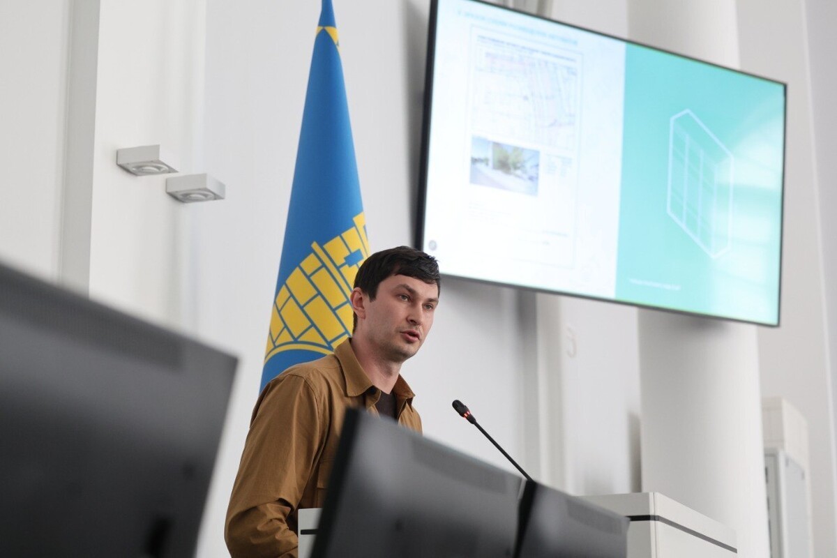 Роман Крушельницький за трибуною у Великій сесійній залі Львівської міськради розповідає підприємцям деталі щодо стандарту автоматів в місті. Позаду нього прапор з Гербом Львова. На стіні екран з презентацією.