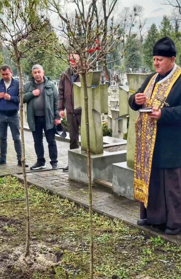 На Меморіалі Січових Стрільців на Янівському кладовищі священник проводить панахиду. На передньому плані видно два щойно висаджені саджанці калини, а біля поховань стоять троє чоловіків - учасників заходу.